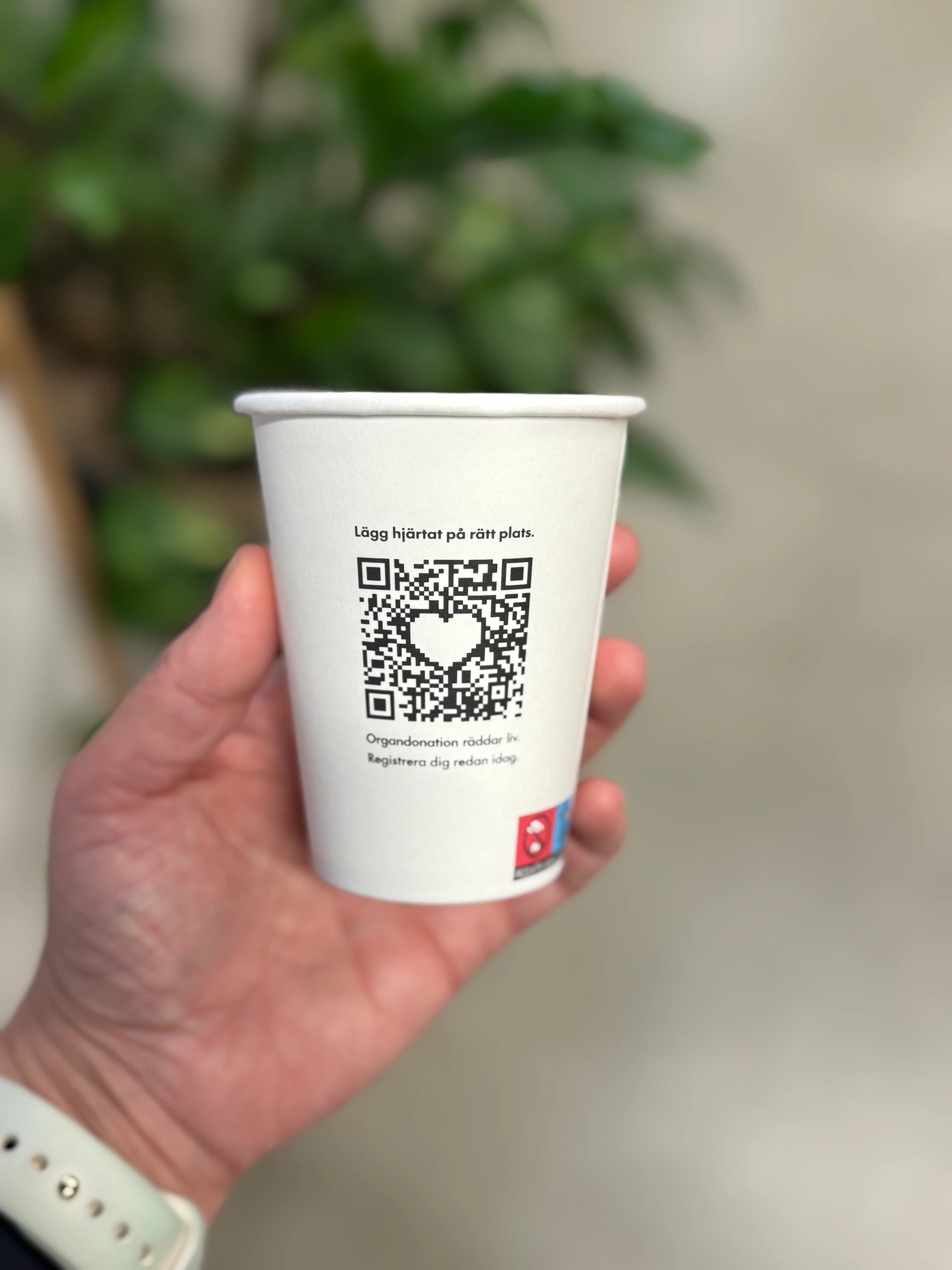 The reverse of the printed Berghs cup, held in a hand, showing the heart-shaped donation-registration QR code and Swedish call to action.